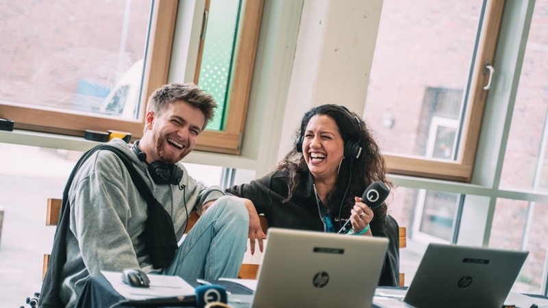 Zwei Mitarbeitende von Radio Bremen sitzen vor Laptops und lachen