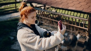 Frau hat ein Handy und ein Mikrofon in der Hand, im Hintergrund sind Tiere zu sehen