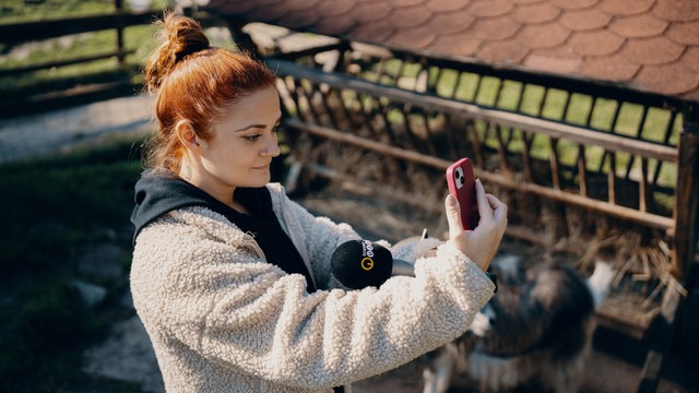 Frau hat ein Handy und ein Mikrofon in der Hand, im Hintergrund sind Tiere zu sehen