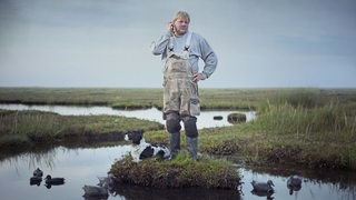 Filmszene: Ein blonder Mann in Arbeitshose steht auf einer kleinen Grasinsel im Moor und kratzt sich am Ohr