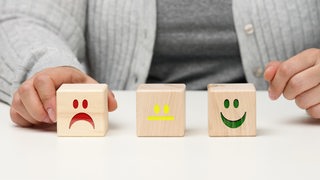 Eine Person sitzt vor 3 Holzwürfeln auf denen Smileys angezeigt werden, die unterschiedliche Stimmungen ausdrücken.