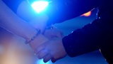 Die Arme eines Mannes bekommen Handschellen angelegt, im Hintergrund leuchtet Blaulicht.
