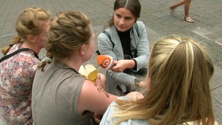 Eine Reporterin sitzt mit Passanten auf einem Fußweg bei einem Interview.