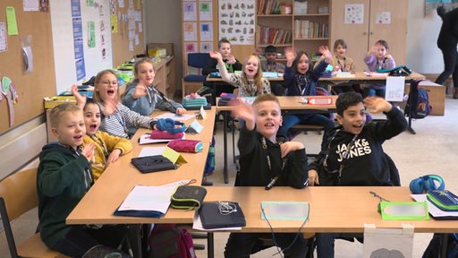 Schülerinnen und Schüler sitzen in einem Klassenzimmer und winken