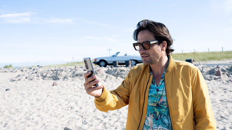 Szenenfoto Oliver Mommsen als Tim Seebach am Strand, dahinter ein Cabriolet
