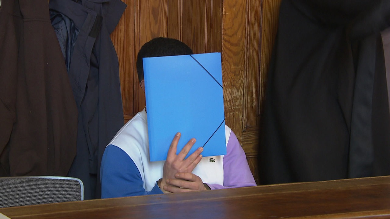 Ein Angeklagter im weißen Pullover sitzt in einem Gerichtssaal, eine blaue Mappe vor dem Gesicht