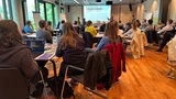 Menschen sitzen in einem Sitzungssaal und schauen auf eine Präsentation