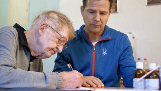 Filmszene der Doku "Dem Sterben zum Trotz", ein alter Mann unterschreibt ein Dokument, neben ihm steht eine Pflegekraft
