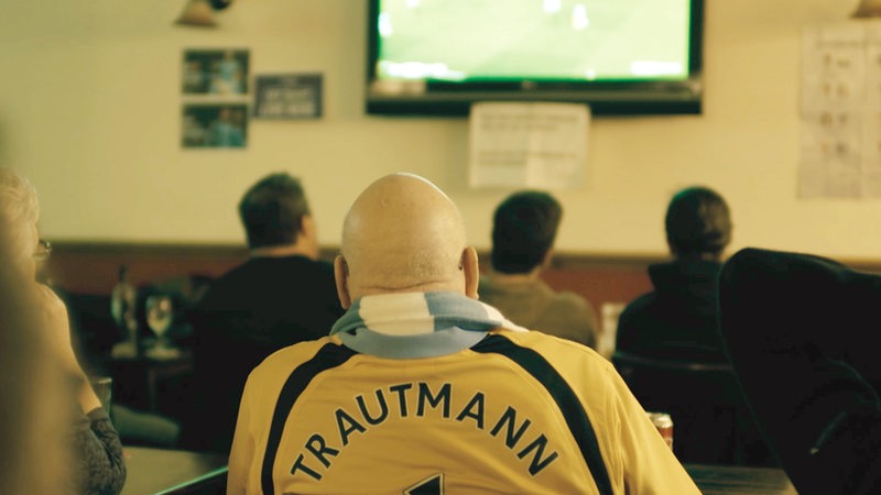 Ein glatzköpfiger englischer Fußballfan mit Trautmann-Trikot sitzt im Pub vor dem Fernseher